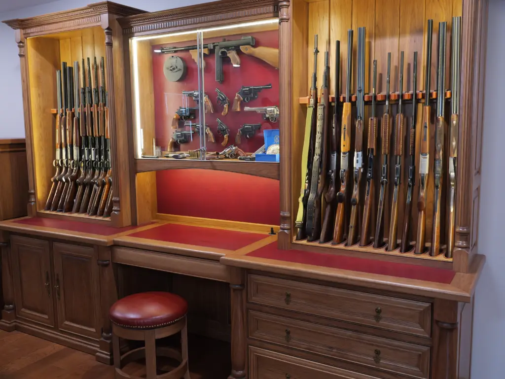 Custom display safe featuring an array of firearms, including rifles and handguns, elegantly showcased in a wooden cabinet with integrated lighting and red accents, emphasizing security and aesthetic appeal for firearm collectors.
