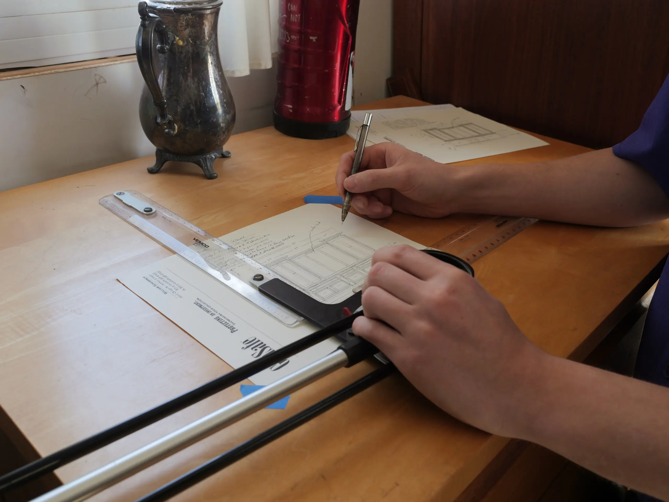 Person measuring and sketching plans for a custom wood gun safe on a wooden desk with a ruler and pen, surrounded by drafting materials and a vintage pitcher.