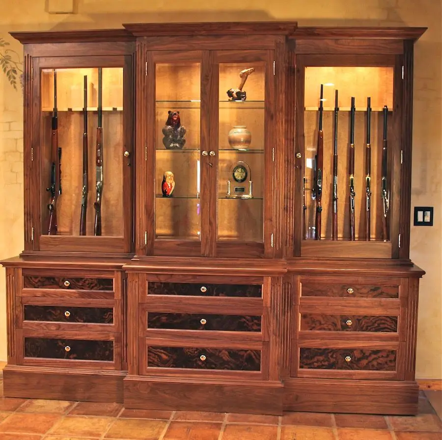 A beautifully lit home study featuring a Custom Cabinet Security handcrafted gun display cabinet with glass doors, LED interior lighting, and rifles and handguns arranged elegantly inside. The room feels warm, responsible, and aspirational. Wide-angle shot, natural and LED light blend.